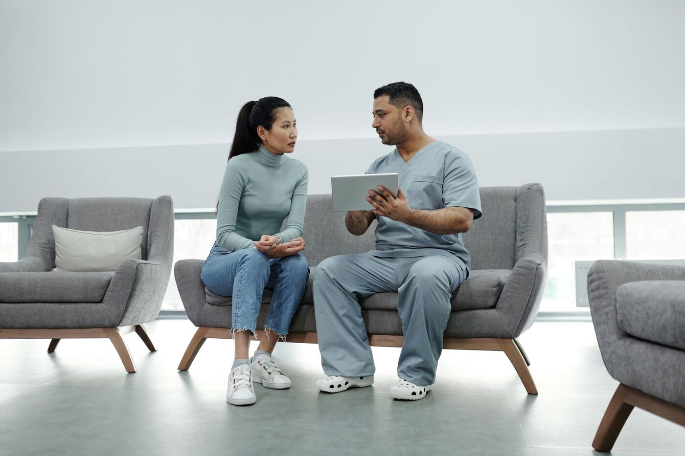 A clinician in scrubs shows a patient a tablet as they sit together on a sofa.