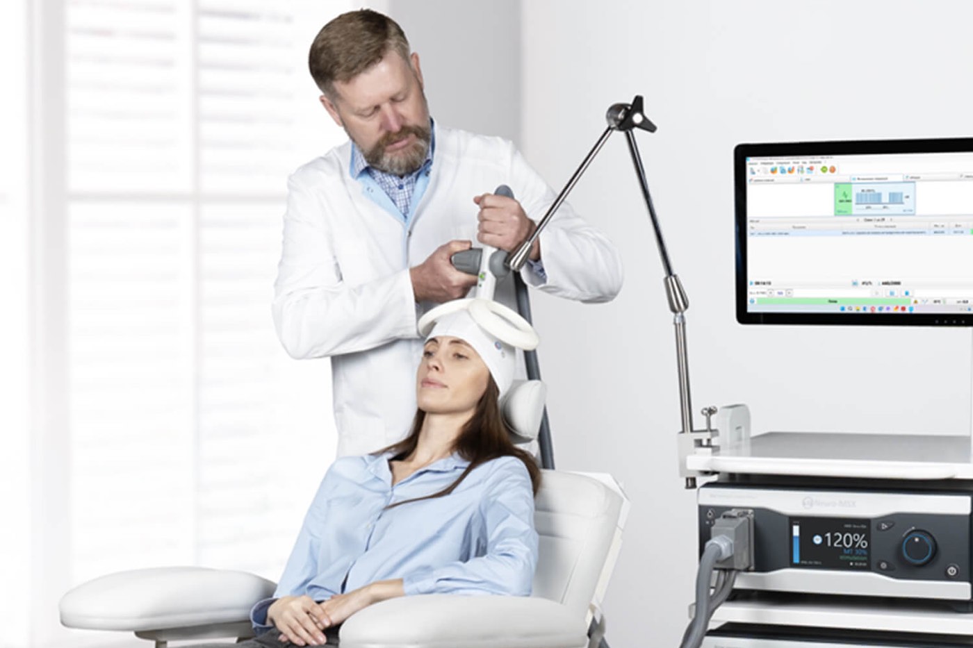 A clinician positions a TMS coil on a seated patient, with the stimulator and its treatment screen beside them.